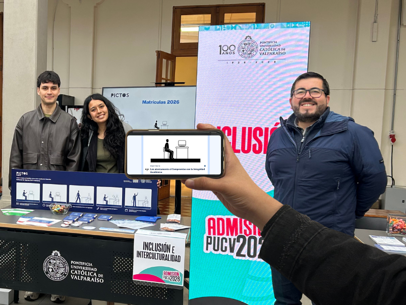 Equipo profesional en stand Dirección de Inclusión. En parte frontal mano sosteniendo un celular con gráfica de PICTOS.