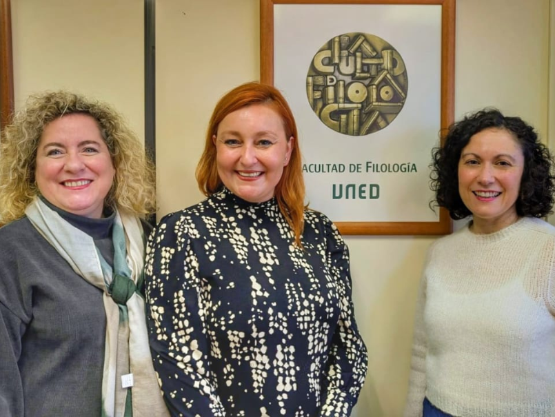 Jadranka Gladic sonríe en fotografía junto a dos profesionales de universidad española. Atrás placa Facultad de Filología UNED. 